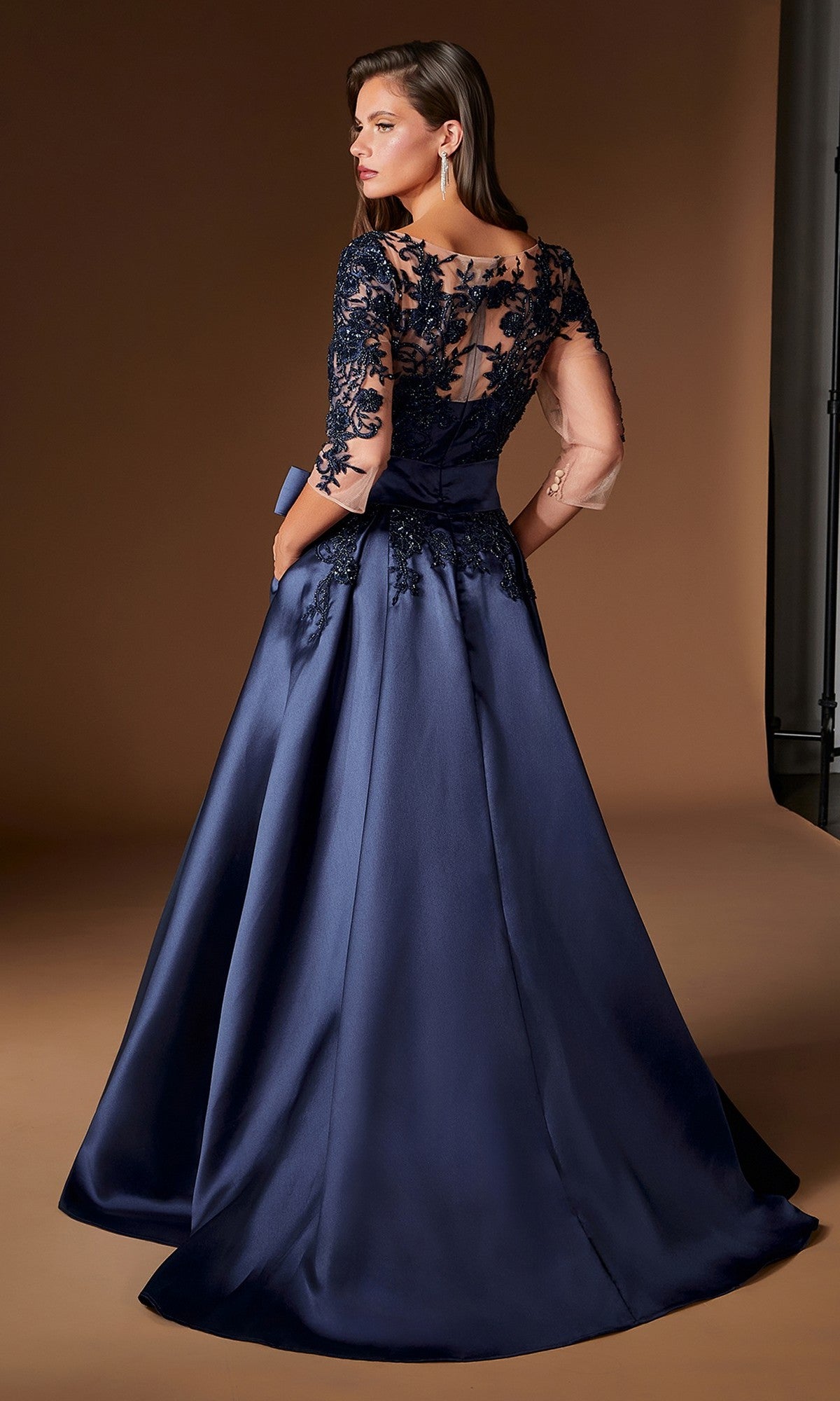 A woman wearing the Ladivine CDS569 long formal dress, featuring a dark blue, floor-length design with sheer lace sleeves and back, stands against a plain brown backdrop, looking over her shoulder.
